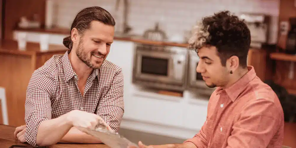 Two individuals sitting at a kitchen table reviewing paperwork together, discussing rental terms or lease agreement.