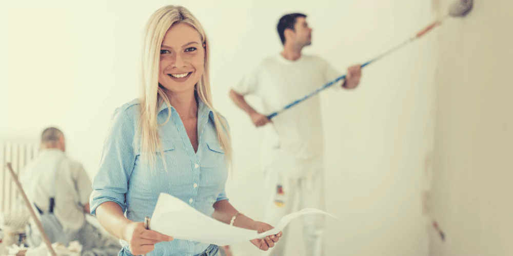 Smiling property manager overseeing interior maintenance as painters work on a rental property wall.
