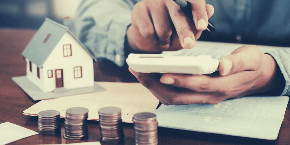 Person calculating property finances with stacked coins, paperwork, and a model house representing rental income management.